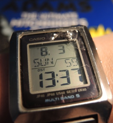 A Casio digital watch displaying Sunday, 3 August, at 13:37:59 daylight savings time, set against a backdrop of The Ultimate Hitchhikers' Guide