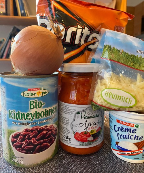 A bag of Doritos behind a can of kidney beans, a jar of Ajvar, some crème fraîche, a bag of grated cheese, and an onion.