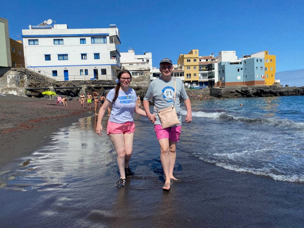Angela and Joey holding hands walking in wet sand along the edge of the waves on a beach in Tenerife. We're both wearing pink board shorts and light-coloured T-shirts. In the background are blue skies and apartment buildings.