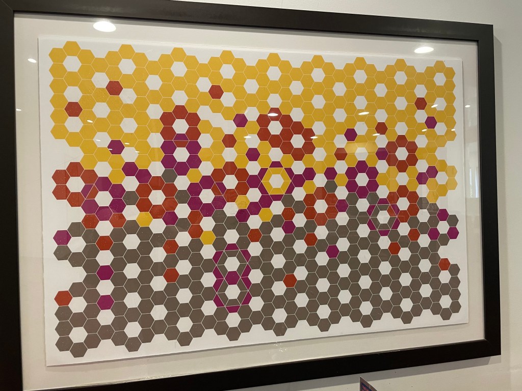 A framed abstract artwork made of rings of coloured rings of hexagons each with a white hexagon in the middle. On the top half the hexagons are mostly yellow, and on the bottom half they're mostly grey, but in between there are some red and purple rings. Sometimes, where the rings meet, the hexagons are split in through the middle, with a different colour on each side. There are also occasional lone red or purple hexagons as part of yellow or grey rings.