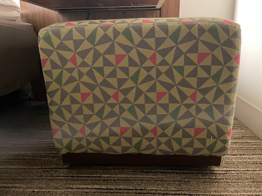 A couch patterned with rectangles that are each divided through the centre into either four or two triangles (with the two-triangle rectangles being in groups of four so they could be seen as an eight-triangle square), with most triangles being grey or off-white, but some in pink or green. 