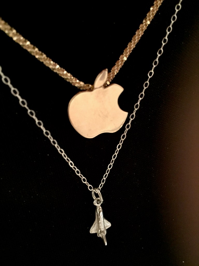 A gold Apple logo pendant with no significant scratches or blemishes, hanging on a gold chain above a silver Space Shuttle pendant in similarly good condition on a silver chain.