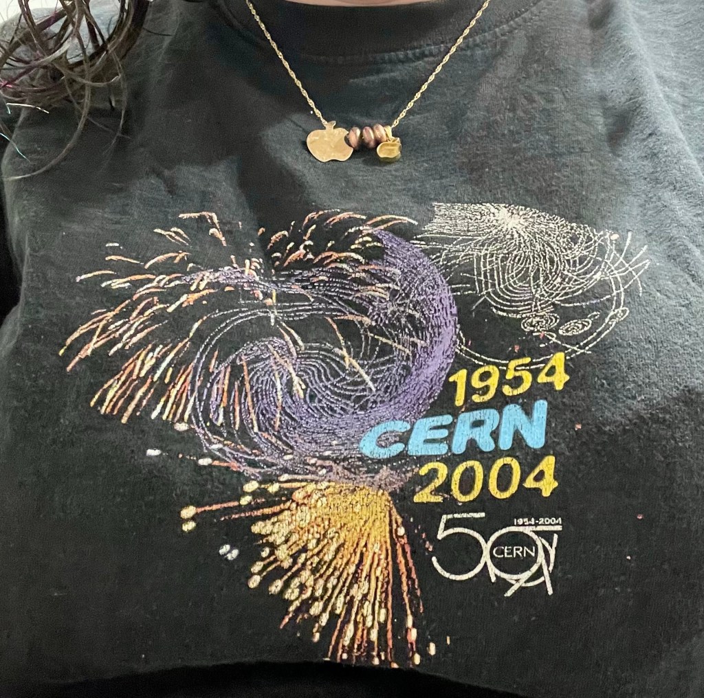 Me wearing a T-shirt that says ‘1954 CERN 2004’ with the CERN 50th anniversary logo (50 where the 0 looks like an accelerator complex) and a design featuring fireworks and particle tracks. I’m also wearing a gold chain with two different gold Apple logo pendants, separated by three pinkish gold beads.